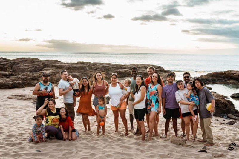 A large multiracial group of adults and children stand on a sandy beach in Hawaii at sunset and smile directly at the camera.